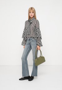 Melnā un baltā šaha raksta blūze ar zvana piedurknēm, pārī ar gaiši zilām platiām džinsēm un zaļu rokassomu. Modelis stāv uz vienkārša fona.