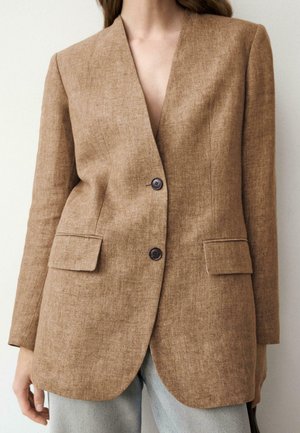 Femme portant un blazer marron clair à deux boutons avec poches à rabat sur un jean bleu clair, sur un fond clair uni.