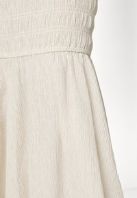 Vestido creme com um tecido texturizado e enrugado. Apresenta um corpete franzido e uma saia rodada, destacando um design leve e suave.