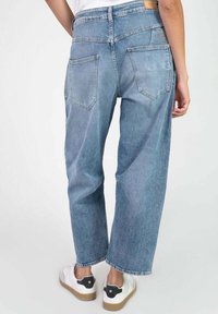 Une personne portant un jean en denim bleu clair à coupe décontractée et des baskets blanches avec des accents noirs au niveau du talon, montrée de dos sur un fond uni.