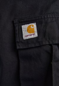 Black fabric patch with white stitching and a rectangular label featuring a yellow Carhartt logo and maroon text. Durable cotton material.