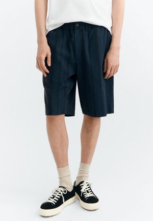 Person trägt schwarze, gestreifte Shorts bis zum Knie, beige Socken, schwarz-weiße Schnürschuhe, mit entspannt an den Seiten hängenden Händen.