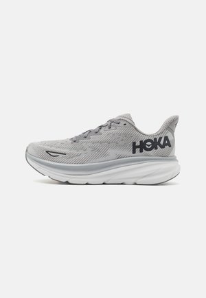 Hellgrauer sportlicher Laufschuh mit großem schwarzem "HOKA"-Logo an der Seite und dicker weißer, strukturierter Sohle, von der Außenseite betrachtet.