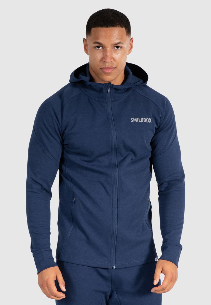 Felpa con zip blu navy realizzata in tessuto liscio, dotata di cappuccio, maniche a raglan e dettaglio del logo sul petto. Resistente e con tasche laterali.