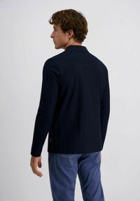 Marineblaue Langarmbluse mit strukturiertem Kragen, aus einem texturierten Stoff und im taillierten Schnitt. Getragen mit hellblauen Hosen.