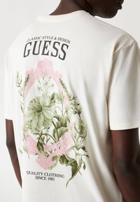 Camiseta de manga corta color crema con un gráfico floral en verde y rosa en la parte posterior, que presenta el texto "GUESS" y "ROPA DE CALIDAD DESDE 1981."
