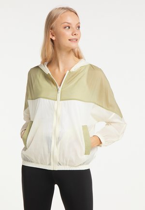 Junge Frau trägt eine hellgrüne und weiße Kapuzenjacke mit Reißverschluss, hat die Hände in den Taschen, schaut seitlich und lächelt.