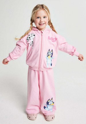 Young girl with blonde braids wearing a pink Bluey-themed tracksuit and light pink shoes, standing with arms outstretched.