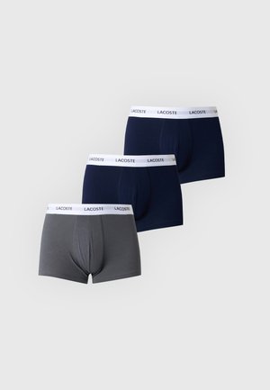 Homme athlétique debout pieds nus portant un boxer noir Lacoste avec une ceinture blanche sur un fond clair uni.