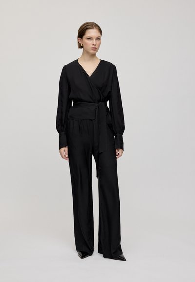 Zwarte wikkelblouse met lange mouwen en een strikceintuur, gecombineerd met loszittende zwarte broek. Glad, glanzend materiaal met minimale details.