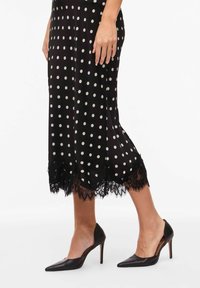 Jupe noire à pois avec un bord en dentelle à l'ourlet, associée à des talons pointus noirs, mettant en avant un design élégant et une texture lisse.