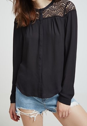 Czarna bluzka z długim rękawem z koronkowymi zdobieniami na ramionach, zapinana z przodu na guziki, noszona z jasnoniebieskimi, postrzępionymi szortami z denimu.