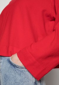 Pull en maille rouge avec une coupe décontractée et des manches longues, associé à un jean en denim bleu clair, montrant une main dans la poche.