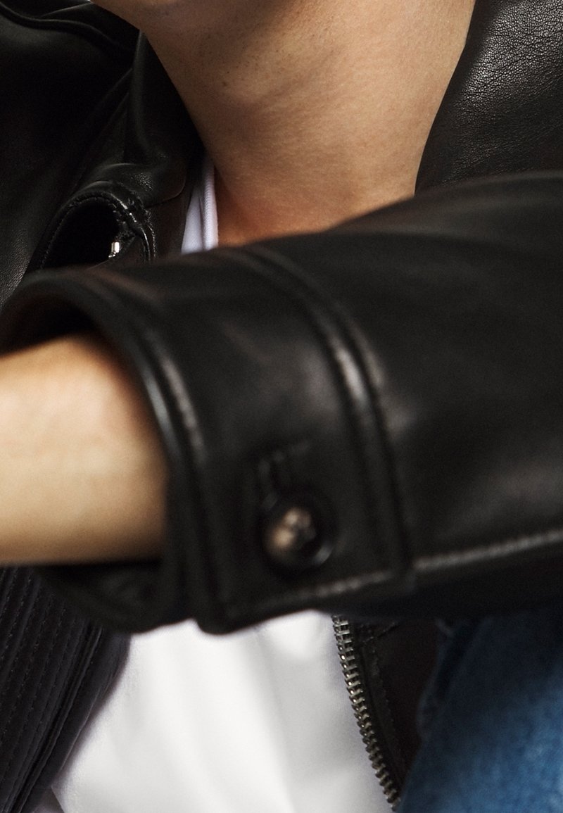 Gros plan d'une personne portant une veste en cuir noire par-dessus une chemise blanche, montrant les détails du col et des manches.