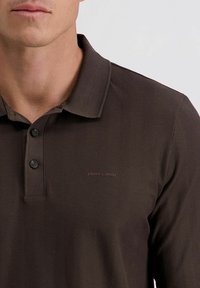 Braunes Poloshirt mit einem Fischgrätenmuster, dreiknöpfiger Knopfleiste und dezentem Branding auf der Brust. Der Stoff wirkt strukturiert und glatt.