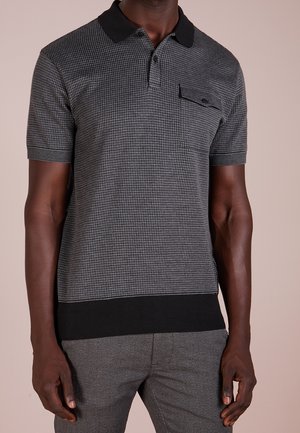 Homme portant un polo texturé gris et noir à manches courtes avec une poche poitrine boutonnée et un pantalon gris sur un fond uni.