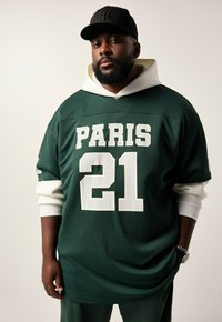 Jersey verde con estampado blanco "PARIS 21". Presenta un diseño con capucha y una capa interior blanca. Se lleva con pantalones verdes y una gorra negra.