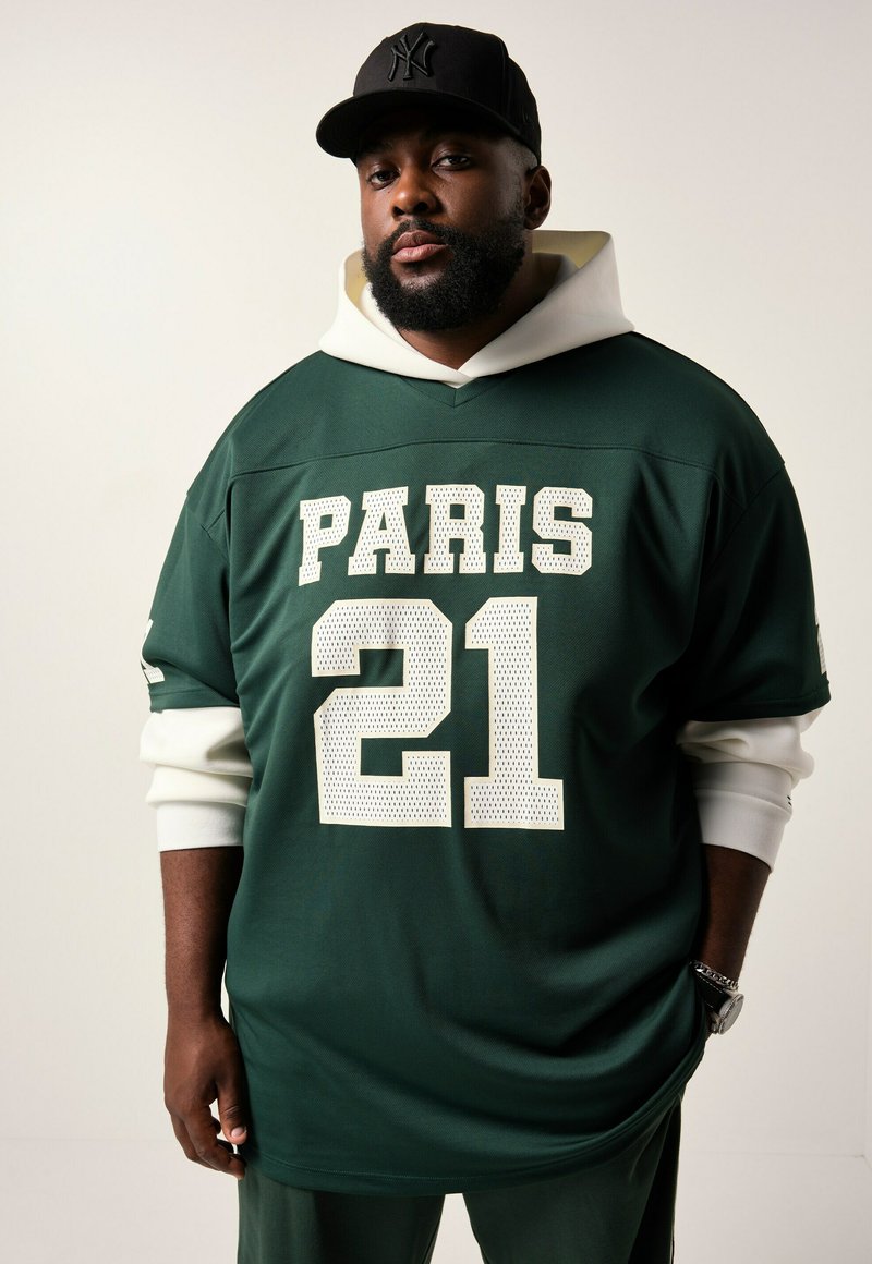 Jersey verde con estampado blanco "PARIS 21". Presenta un diseño con capucha y una capa interior blanca. Se lleva con pantalones verdes y una gorra negra.