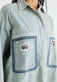 Camicia di denim a righe in blu chiaro e bianco, con due tasche a toppa bordate di blu in contrasto, bottoni in metallo e un'etichetta con logo.