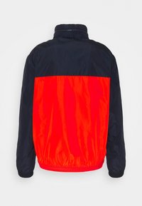 Punainen ja laivastonsininen tuulitakki, jossa on korkea kaulus, joustava helma ja hihnat. Ominaisuuksiin kuuluu kaksivärinen muotoilu ja sileä, kiiltävä pinta.