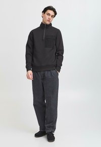 Hanorac negru cu fermoar, prevăzut cu o buzunar frontal, purtat cu blugi gri închis, largi, și încălțăminte neagră. Material neted și design minimalist.