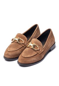 Mocassini in suede marrone con suola piatta nera, caratterizzati da un accento in metallo dorato sulla tomaia e dettagli cuciti sulla parte superiore.