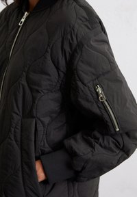 Veste bomber matelassée noire avec des poignets côtelés, dotée d'une poche latérale et d'une poche zippée sur le bras. Tissu lisse et léger avec une finition texturée.