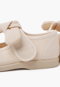 Beige Leinenschuhe mit runder Zehenpartie, Knoten-Detail am Riemen und einer weichen Gummisohle. Strukturierter Stoff mit sichtbaren Nähten.