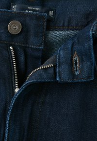 Jean en denim bleu foncé avec une fermeture éclair argentée partiellement ouverte, un bouton métallique portant l'inscription "MNG Denim" et un boutonnière cousue visible.