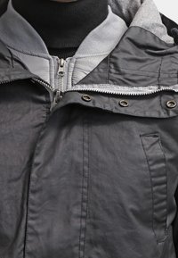 Gros plan sur une veste grise à couches avec capuche, fermeture éclair, boutons-pression, et un col roulé noir en dessous.