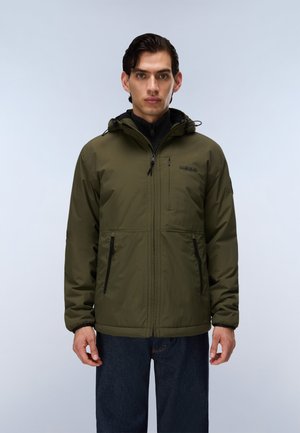 Mann trägt eine olivgrüne Jacke mit Reißverschluss und Kapuze sowie schwarze Reißverschlusstaschen, steht vor einem einfarbigen hellen Hintergrund.