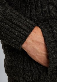 Mørkegrå strikket sweater med ribbet tekstur, hvor en hånd er placeret i sidelommen, hvilket fremhæver stoffets tykkelse og blødhed.