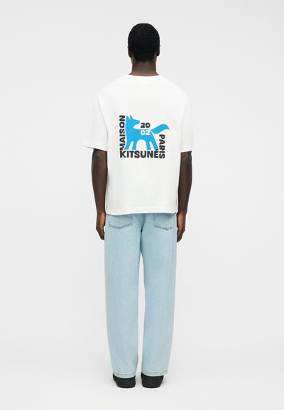 Άνδρας που φοράει λευκό μπλουζάκι με μπλε αλεπού και μαύρο κείμενο «Maison Kitsuné Paris 2002» στην πλάτη, ανοιχτό μπλε τζιν και μαύρα παπούτσια, όρθιος.