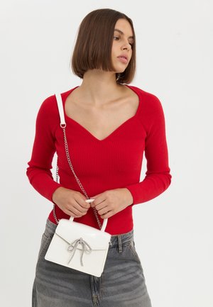 Femme aux cheveux courts bruns portant un haut à manches longues rouge et un jean gris, tenant un sac à main blanc avec un détail de nœud argenté.