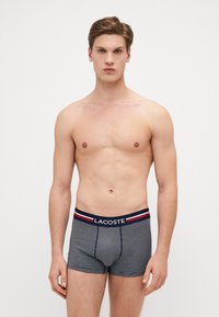 Lacoste Herren Boxershorts in Marineblau mit weißen Streifen. Der elastische Bund hat ein Logo und rote Akzente. Glatter Stoff und enge Passform.