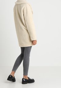 Persona con un cappotto teddy beige lungo fino al ginocchio, jeans skinny grigi e mocassini neri spessi in piedi contro uno sfondo bianco semplice.
