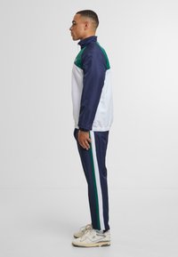 Tuta sportiva con una combinazione di colori blu navy e bianco, con accenti verdi sulle spalle e strisce laterali. Realizzata in materiale leggero.