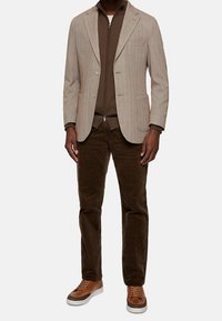 Blazer de patrón espiga en marrón claro, combinado con un suéter marrón de cremallera, pantalones de pana oliva y zapatillas marrones.