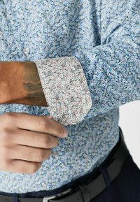 Ziedu raksta krekls gaiši zilā audumā ar garām piedurknēm, kam ir kontrastējoša aproces raksts ar daudzkrāsainām ziedu motīviem un pogu aizdari.