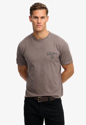 Mand med kort hår iført en grå Superdry t-shirt og sorte bukser, stående med hænderne bag ryggen mod en ensfarvet baggrund.