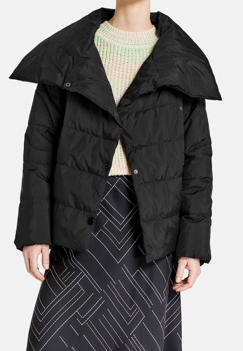 Svart puffjacka med stor krage, kviltad textur och knäppning, kombinerad med en flerfärgad stickad tröja och mönstrad kjol.