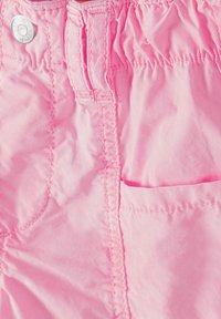 Gros plan sur un tissu rose montrant un bouton en argent, une taille froncée, une passant de ceinture et des coutures de poche sur des vêtements.