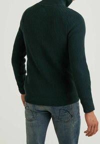 Tmavě zelený žebrovaný svetr s vysokým límcem a dlouhými rukávy, v kombinaci se světle modrými džíny. Viditelné švy a strukturovaná tkanina.