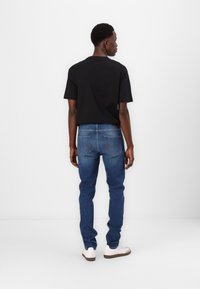 Man wearing a black short-sleeve shirt and blue jeans, with white sneakers. The jeans feature a classic five-pocket design.