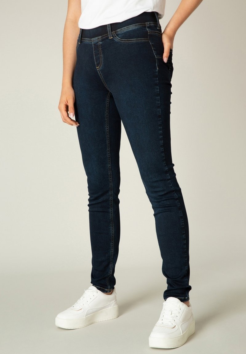 Donkerblauwe skinny jeans gemaakt van denim, met een gladde textuur, contrasterende naden en een middelhoog tailleband. Gecombineerd met witte sneakers.