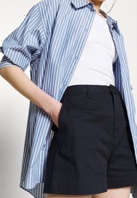 Camisa de rayas azul y blanca de gran tamaño, de mangas cortas, combinada con shorts azul marino. La camisa tiene botones; los shorts tienen un diseño limpio y ajustado.