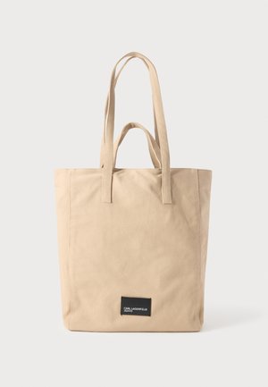 Bež platnena torba s dvije naramenice, s crnom etiketom s bijelim tekstom na dnu za identifikaciju marke.