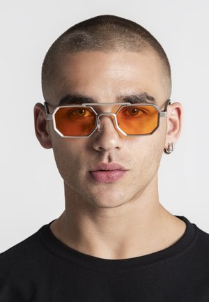 Ung mand med kortklippet hår iført sølvindfattede, orangefarvede, sekskantede solbriller og en sort skjorte foran en ensartet lys baggrund.