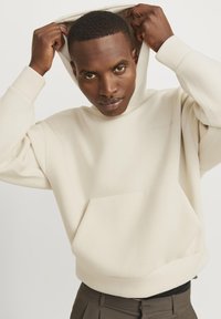 Roomkleurige hoodie van zacht materiaal met een voorzak, een capuchon met trekkoord en minimalistische branding op de borst.