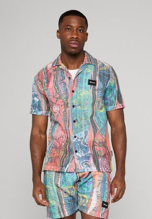 Mies, joka pukeutunut värikkääseen, kuviolliseen lyhythihaiseen nappipaitaan ja siihen sopiviin shortseihin yksiväristä taustaa vasten.